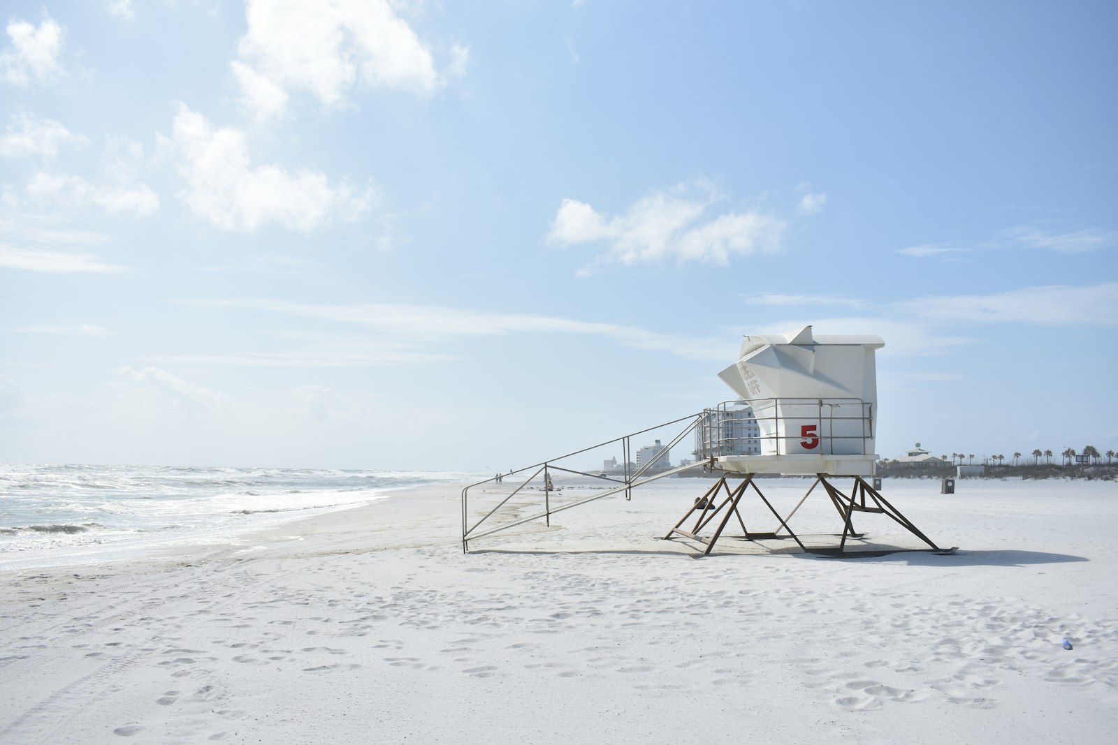 An empty stretch of flat white sand beside a calm sea, with an unmanned lifeguard tower and palms in the distance