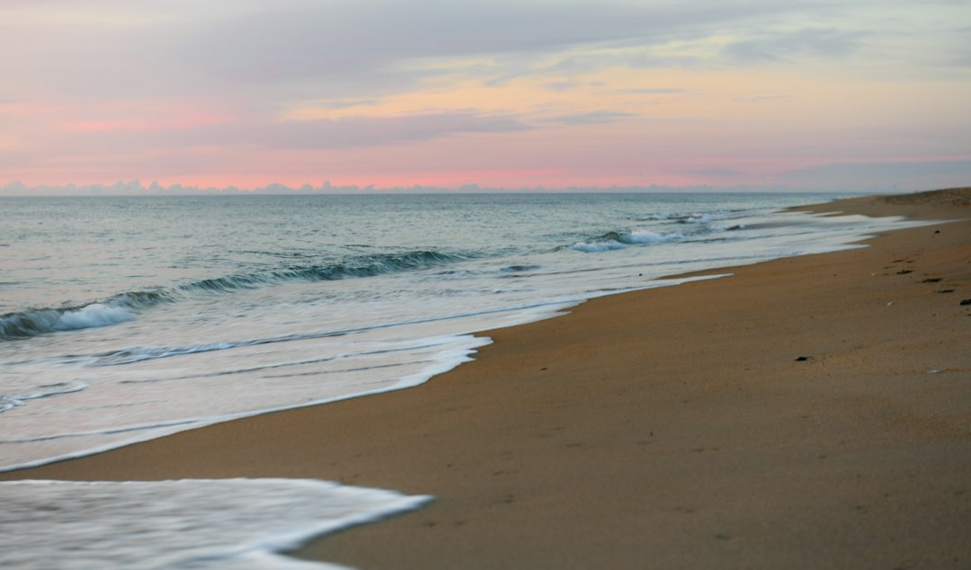 Gentle surf rolling onto a wide, flat sand beach at Kill Devil Hills under a pastel sunset sky